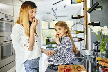 Mother and daughter eating strawberries in modern kitchen - ZEDF02339