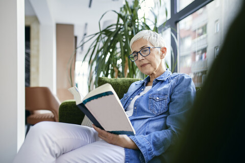 Ältere Frau sitzt auf der Couch und liest ein Buch, lizenzfreies Stockfoto