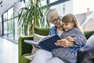 Grandmother sitting on couch with granddaughter, reading book together - ZEDF02368