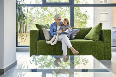 Grandmother sitting on couch with granddaughter, reading book together - ZEDF02369