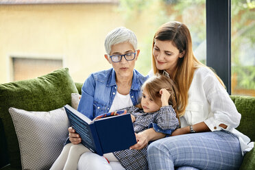 Mother, daughter and granddaughter sitting on couch, reading a book - ZEDF02372