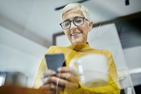 Ältere Frau trinkt Kaffee und überprüft Textnachrichten auf ihrem Smartphone, lizenzfreies Stockfoto