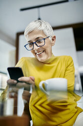 Ältere Frau trinkt Kaffee und überprüft Textnachrichten auf ihrem Smartphone - ZEDF02407