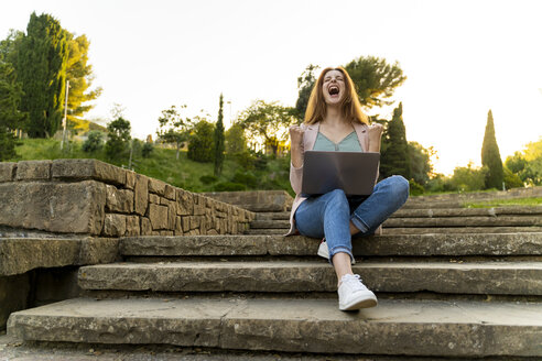 Junge rothaarige Frau, die einen Laptop benutzt und jubelt, sitzt auf einer Treppe in einem Park - AFVF03196