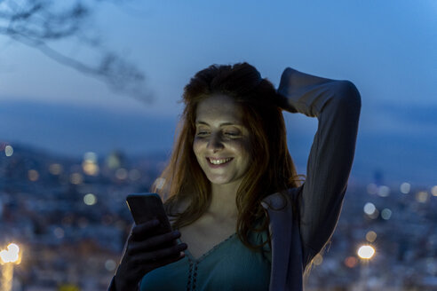 Junge Frau mit Smartphone an einem Aussichtspunkt zur blauen Stunde, Barcelona, Spanien - AFVF03236