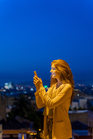 Junge Frau mit Smartphone an einem Aussichtspunkt zur blauen Stunde, Barcelona, Spanien, lizenzfreies Stockfoto