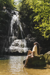 Junge Frau sitzt auf einem Felsen an einem Wasserfall, Garrotxa, Spanien - AFVF03260