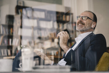 Happy mature businessman listening to music with bluetooth earbuds in a cafe - KNSF05909