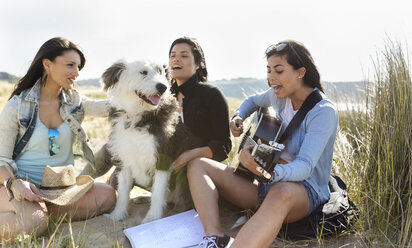 Junge Frau mit Freunden und Hund spielt Gitarre am Strand - MGOF04060