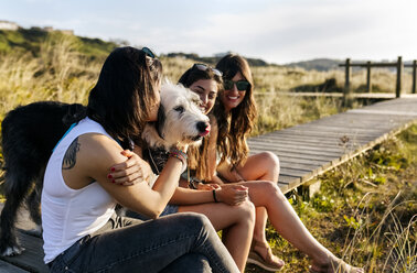 Three happy women with dog sitting on boardwalk in dunes - MGOF04114