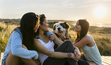 Three happy women with dog sitting on boardwalk in dunes at sunset - MGOF04123