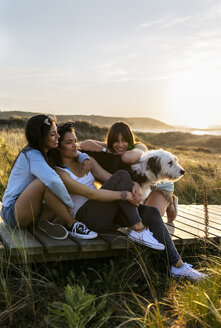 Drei glückliche Frauen mit Hund sitzen auf der Promenade in den Dünen bei Sonnenuntergang - MGOF04124