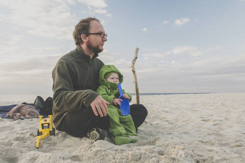Vater sitzt mit seiner kleinen Tochter an einem Sandstrand, Kurische Nehrung, Litauen, lizenzfreies Stockfoto
