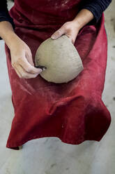 High angle close up of ceramic artist wearing red apron sitting in her workshop, working on clay vase. - MINF11331