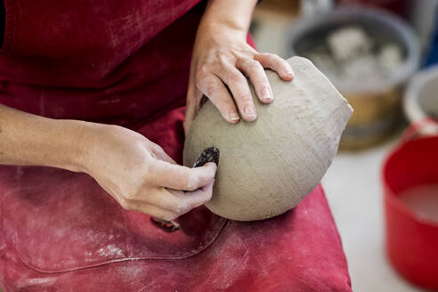 High angle close up of ceramic artist wearing red apron sitting in her workshop, working on clay vase. - MINF11332