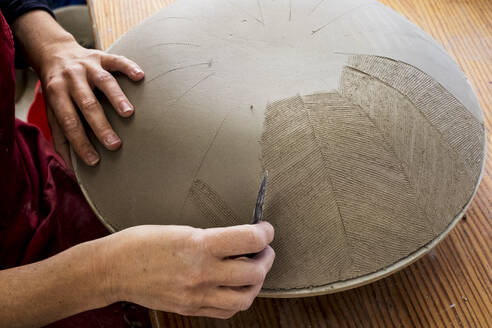 High angle close up of ceramic artist working on clay bowl, applying pattern with hand tool. - MINF11333