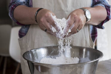 Nahaufnahme einer Person in Schürze, die in der Küche steht und in einer Metallschüssel die Zutaten für einen Streuselkuchen mischt. - MINF11364