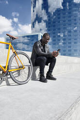 Mann mit gelbem Fahrrad, der auf einer Treppe sitzt und ein Smartphone benutzt, Barcelona, Spanien - JND00056