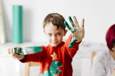 Boy showing a brush in one hand and the other painted green while doing crafts at home - JRFF03242