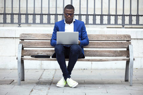 Junger Geschäftsmann mit blauer Anzugsjacke sitzt auf einer Bank und benutzt einen Laptop, lizenzfreies Stockfoto