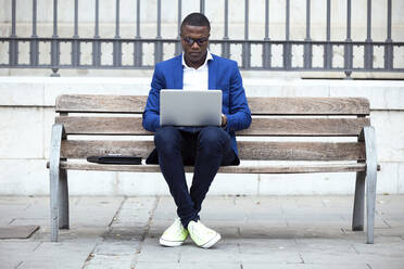 Junger Geschäftsmann mit blauer Anzugsjacke sitzt auf einer Bank und benutzt einen Laptop - JSRF00233