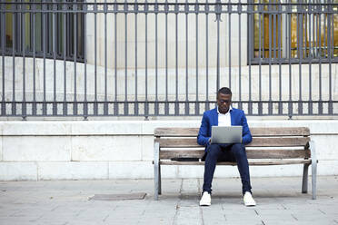 Junger Geschäftsmann mit blauer Anzugsjacke sitzt auf einer Bank und benutzt einen Laptop - JSRF00234