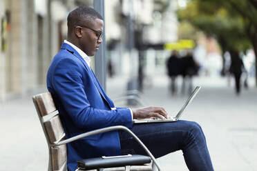 Junger Geschäftsmann mit blauer Anzugsjacke sitzt auf einer Bank und benutzt einen Laptop - JSRF00235