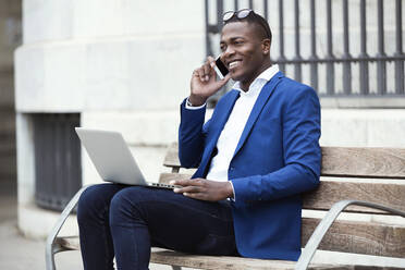 Young businessman wearing blue suit jacket sitting on bench and using smartphone - JSRF00237