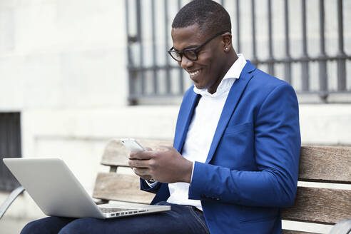 Junger Geschäftsmann mit blauer Anzugsjacke sitzt auf einer Bank und benutzt ein Smartphone - JSRF00240