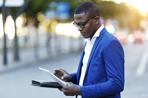 Junger Geschäftsmann in blauer Anzugsjacke, der sein digitales Tablet auf einer Straße benutzt, lizenzfreies Stockfoto