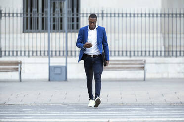 Junger Geschäftsmann mit blauer Anzugsjacke, der die Straße überquert und ein Smartphone benutzt - JSRF00257