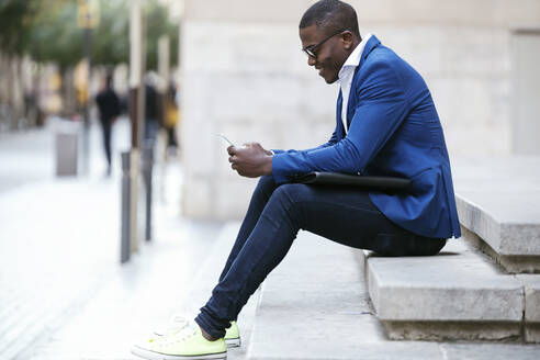 Junger Geschäftsmann mit blauer Anzugsjacke, der auf einer Stufe sitzt und ein Smartphone benutzt - JSRF00259