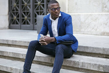 Junger Geschäftsmann mit blauer Anzugsjacke, sitzt auf einer Stufe und hält ein Smartphone in der Hand - JSRF00261