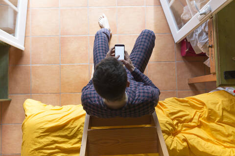 Mann benutzt Smartphone, sitzt auf Holzleiter im Schlafzimmer, lizenzfreies Stockfoto