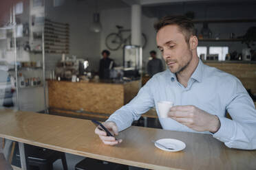 Erfolgreicher Geschäftsmann sitzt in einem Café, trinkt Kaffee und benutzt sein Smartphone - KNSF06019