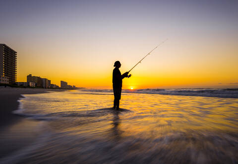 Unscharfe Ansicht der Silhouette eines Mannes beim Fischen in den Wellen am Strand bei Sonnenuntergang, lizenzfreies Stockfoto