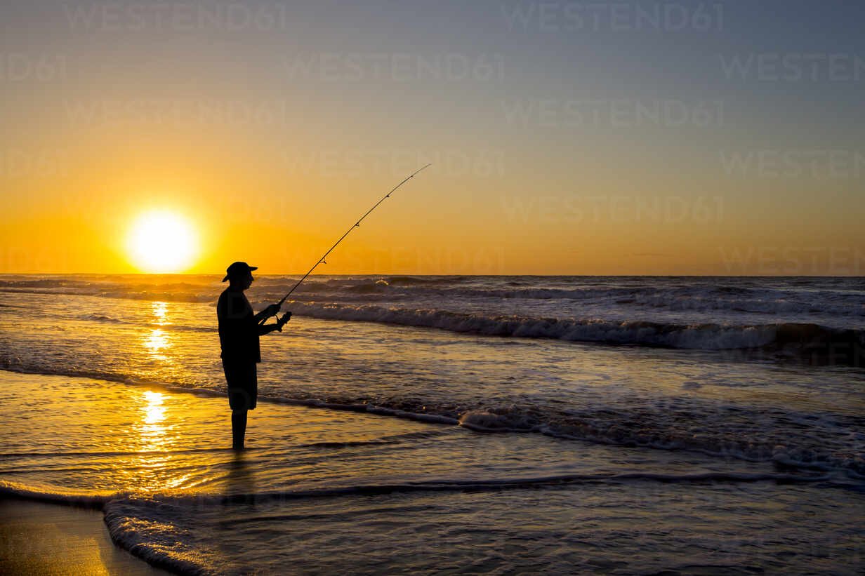 Silhouette of man fishing in waves on beach at sunset stock photo