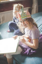 Two sisters wearing crowns and unpacking presents on bed - IHF00107