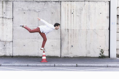 Young man balancing on traffic cone in front of concrete wall - UUF17828