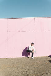 Junger Mann vor rosafarbenem Bauzaun, auf einem Bein hockend - UUF17858