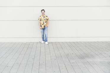 Happy young man wearing aloha shirt, standing against house wall - UUF17864