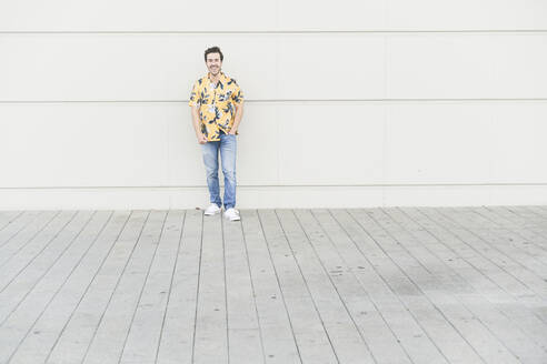 Glücklicher junger Mann mit Aloha-Hemd, der an einer Hauswand steht - UUF17864