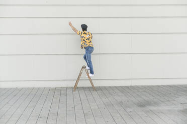 Young man wearing flat hat and aloa shirt, standing on step ladder, touching wall - UUF17877