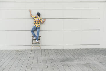 Young man wearing flat hat and aloa shirt, standing on step ladder, touching wall - UUF17878