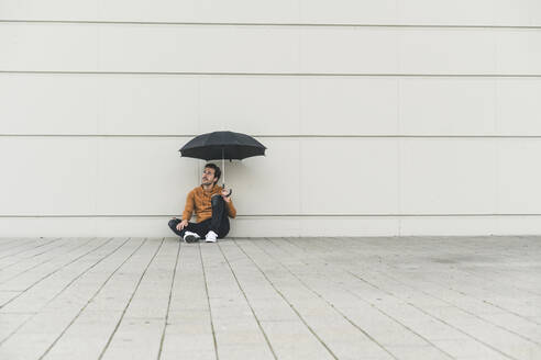 Junger Mann mit Regenschirm, auf dem Boden sitzend, unsicherer Blick - UUF17883