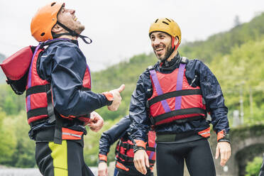 Laughing friends in protective wear at river shore - FBAF00742