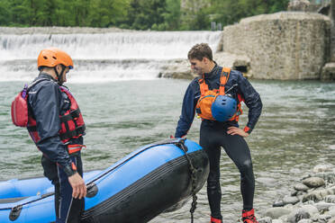 Two men with rubber dinghy at river shore - FBAF00746