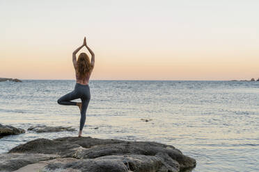 Junge Frau beim Yoga am Strand, in Baumhaltung, während des Sonnenuntergangs an einem ruhigen Strand, Costa Brava, Spanien - AFVF03275