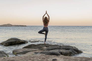 Junge Frau beim Yoga am Strand, in Baumhaltung, während des Sonnenuntergangs an einem ruhigen Strand, Costa Brava, Spanien - AFVF03277