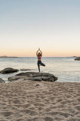 Junge Frau beim Yoga am Strand, in Baumhaltung, während des Sonnenuntergangs an einem ruhigen Strand, Costa Brava, Spanien - AFVF03278